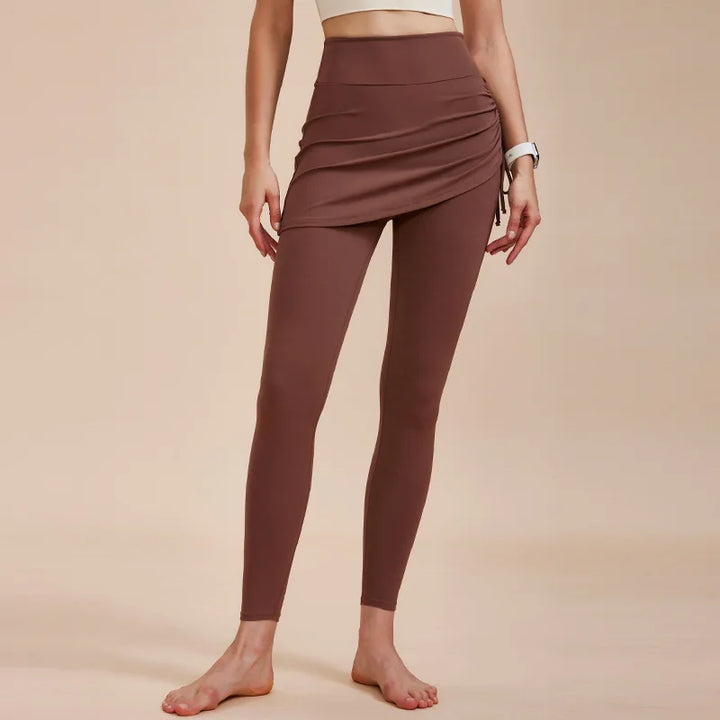 A woman stands barefoot in high-waisted leggings with skirt detailing, paired with a white crop top—an effortlessly chic look for any autumn wardrobe, set against a plain beige background.
