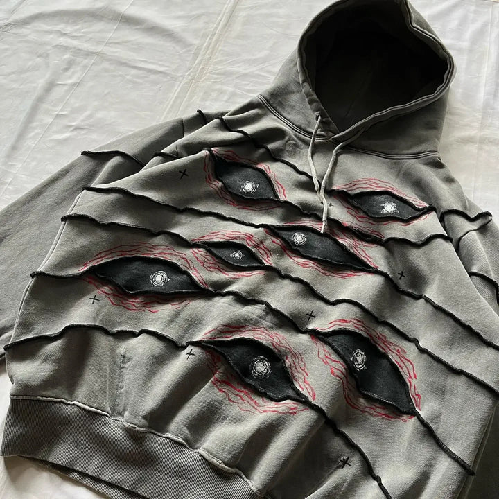 A gray hoodie with a comfortable loose fit, multiple eye designs, and red stitching. Layered, frayed fabric slashes across the front reveal an eye pattern patchwork beneath. The hoodie is laid flat on a white surface.