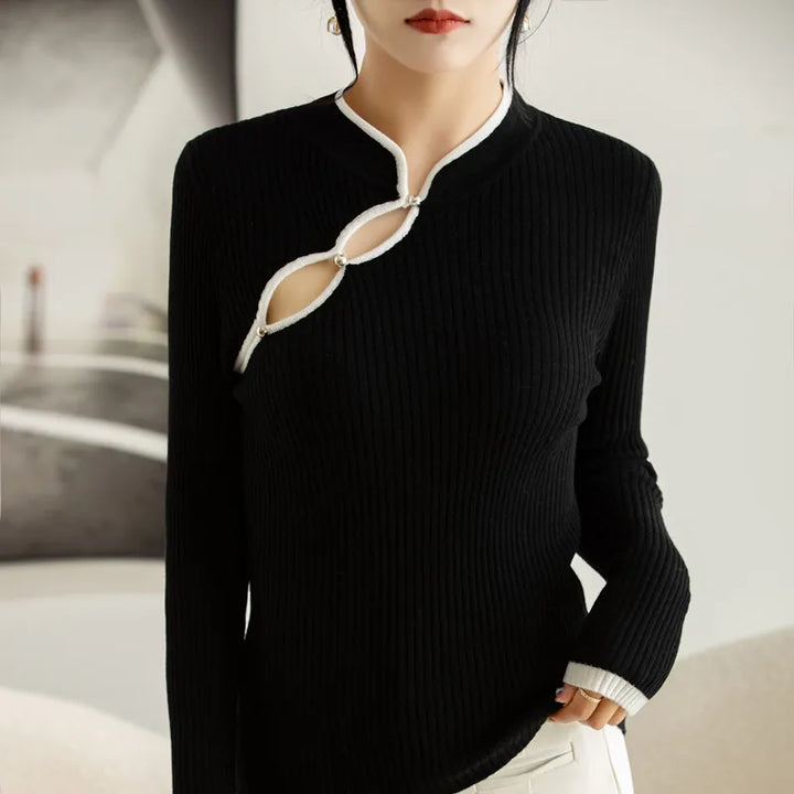 A woman wears a black ribbed knit long-sleeve top with white trim and three keyhole design cutouts along the neckline, paired with white pants. Her face is partially visible and her hand rests in her pocket.