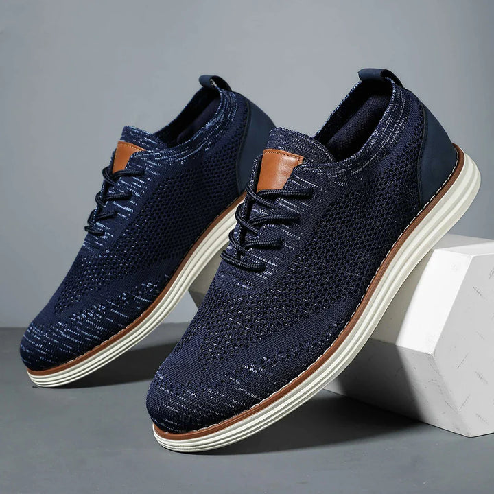 A pair of navy blue breathable knit sneakers with white soles and brown accents, displayed against a gray background. One shoe is elevated on a white block, showing the side and top views for a casual style look.