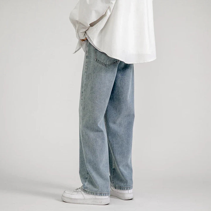 A person wearing light blue premium denim relaxed fit jeans, a white oversized shirt, and white sneakers stands against a plain light gray background, facing sideways with hands partially in pockets.