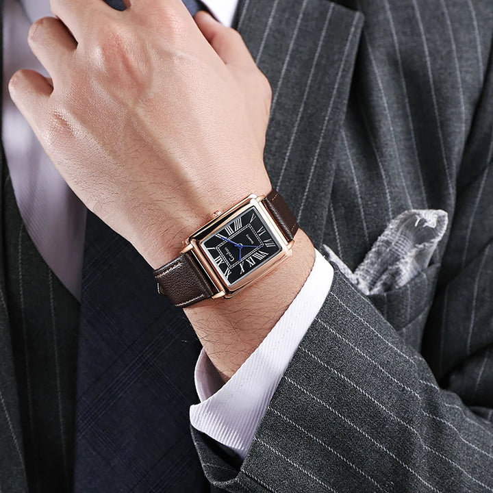 A man wearing a gray pinstripe suit and a patterned tie shows a wrist adorned with a luxury watch featuring a rectangular black face and a premium leather band.