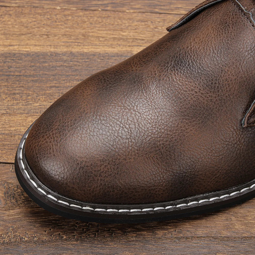Close-up of the front part of an After-Ride Chukka, a brown leather boot with visible texture, white stitching along the black sole, set against a wooden surface background.