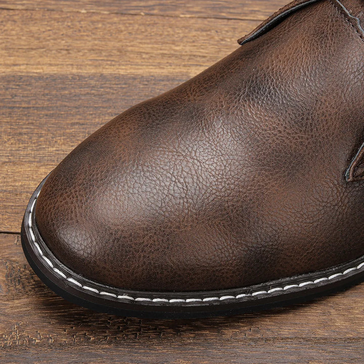 Close-up of the front part of an After-Ride Chukka, a brown leather boot with visible texture, white stitching along the black sole, set against a wooden surface background.
