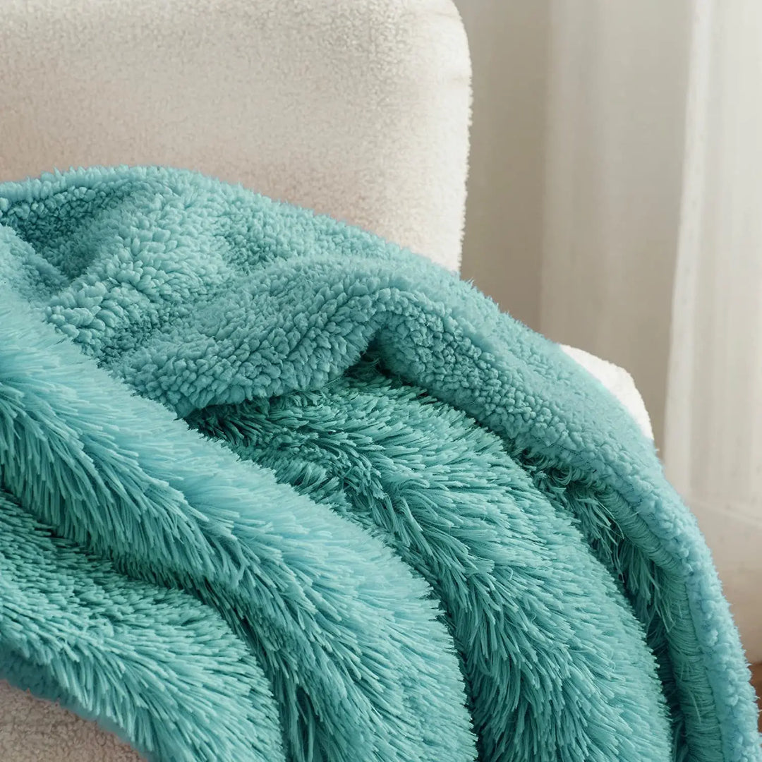 A soft, sage green sherpa blanket with a fluffy texture is draped over the arm of a white upholstered chair, with natural light coming through a window in the background.