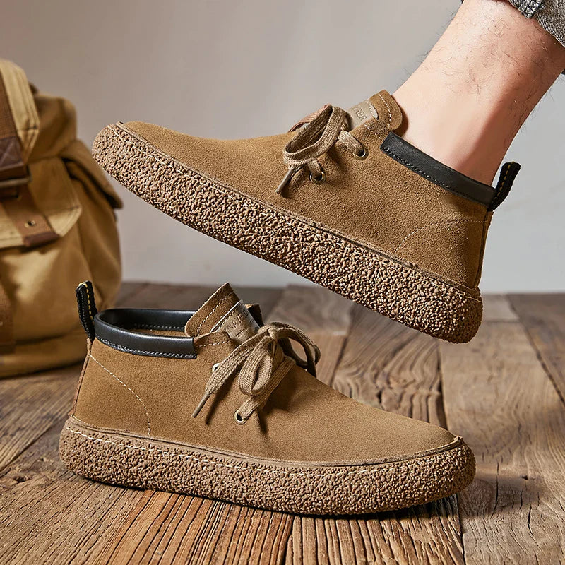 A person wearing brown cow suede leather lace-up sneakers with textured rubber soles stands on a wooden floor. A tan bag is visible in the background.