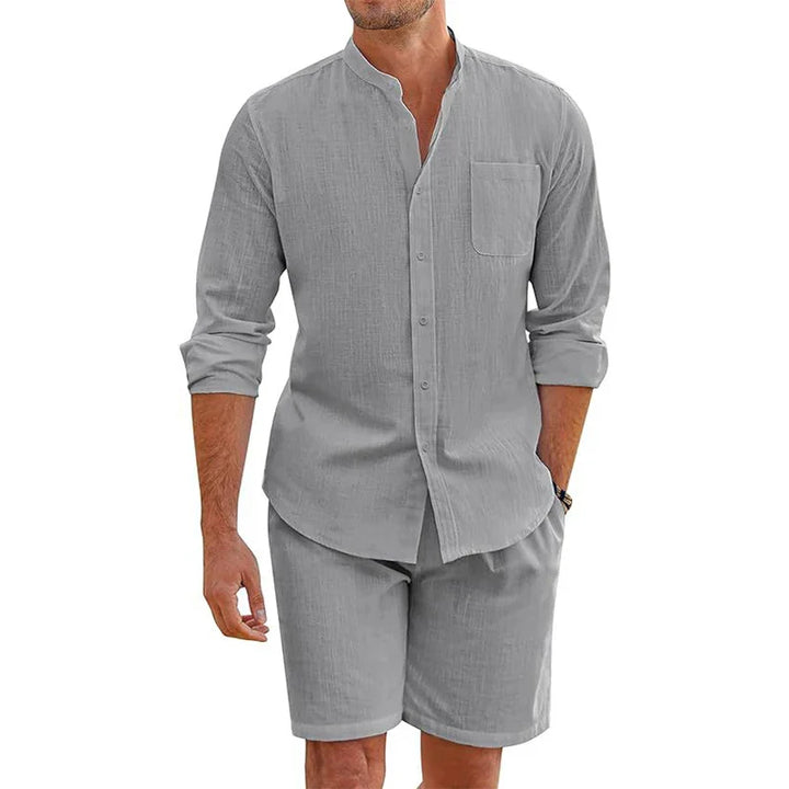 Man wearing a vintage stand collar shirt in light gray cotton linen and matching baggy shorts. One hand is in his pocket while the other arm rests relaxed by his side.
