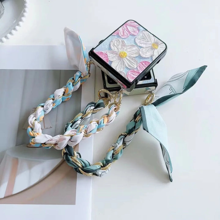 A compact mirror with a floral design and braided pastel handle, adorned with scarf-like fabric and featuring a slim design, placed on a white surface beside a partially visible magazine or photo.