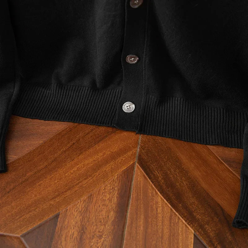 Close-up of the bottom part of a relaxed fit black buttoned cardigan lying on a wooden surface with a geometric pattern. Only the ribbed hem and three buttons are visible.