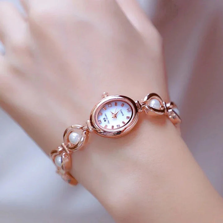 A delicate rose gold wristwatch with a round white face and Roman numerals, featuring precision quartz movement and pearl accents on infinity-shaped links, worn on a persons slender wrist.