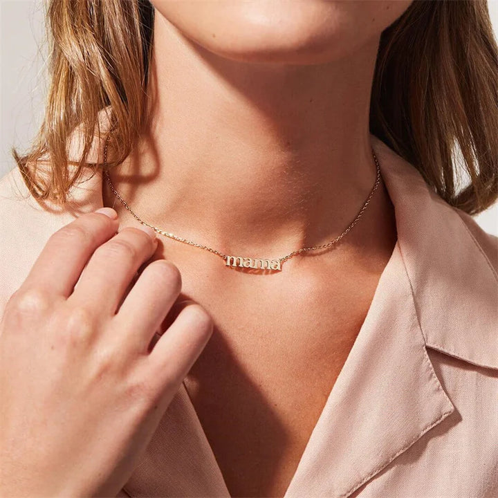 A person wearing a pale pink blouse touches a Mama Forever Necklace, featuring a gold mama pendant spelled in lowercase letters.