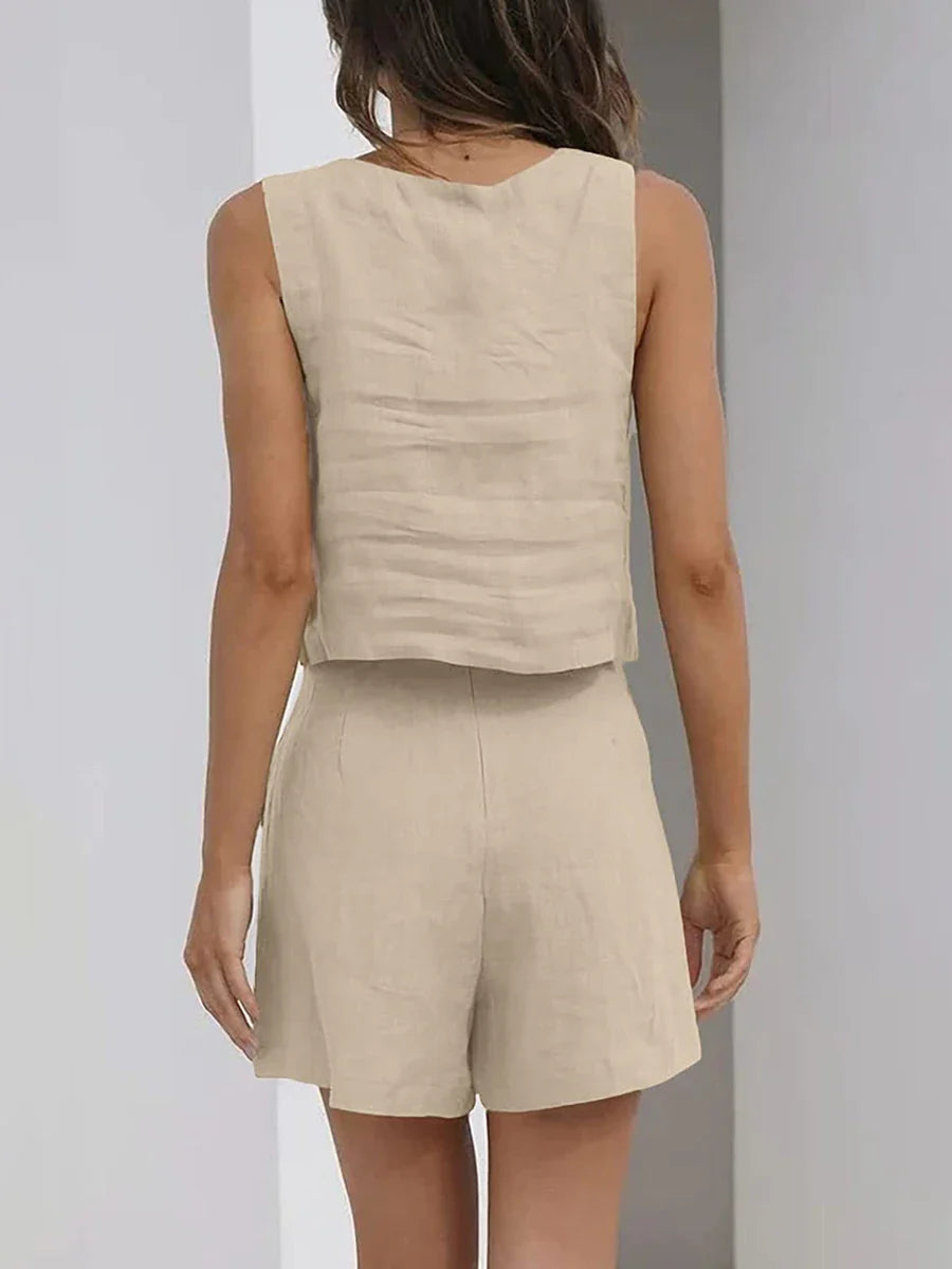 A woman stands indoors facing away, wearing a contemporary style linen set—a sleeveless beige vest and matching high-waisted shorts. Her arms are relaxed at her sides, and her brown hair is loose, partly covering her shoulder.