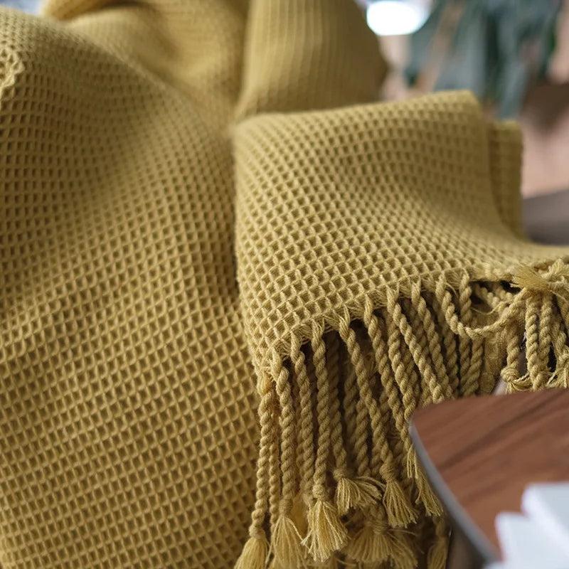 A close-up of a mustard yellow knit waffle blanket with tasseled fringe, draped over a surface. This decorative accent blanket features a textured weave that adds cozy boho charm to any space.
