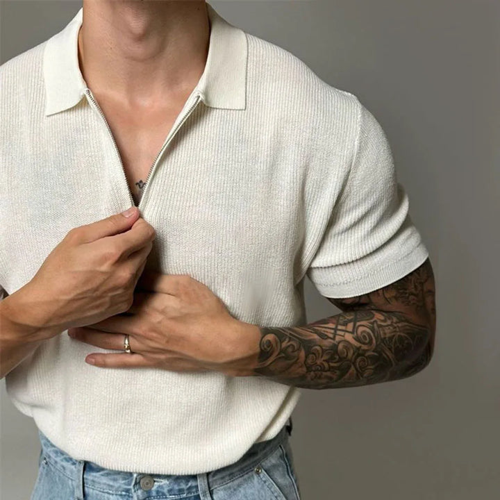 A person wearing a cream-colored quarter zip polo with ribbed detailing and blue jeans, partially unzipping the collar. One hand shows a wedding ring, and the other arm has a detailed sleeve tattoo. The face is not visible.