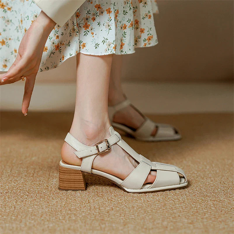 A person wearing a white floral dress and beige heeled sandals with ergonomic arch support stands on a tan carpet, one hand reaching down toward their ankle.
