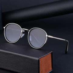 Round, silver-framed eyeglasses with clear, scratch resistant lenses are displayed on a black rectangular case against a dark background. The glasses feature thin metal arms and nose pads for comfort.