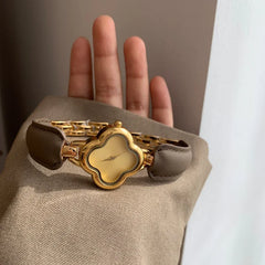 A luxury gold-plated clover dial watch with a genuine leather strap rests on a person’s raised hand, partially covered by a beige sleeve.