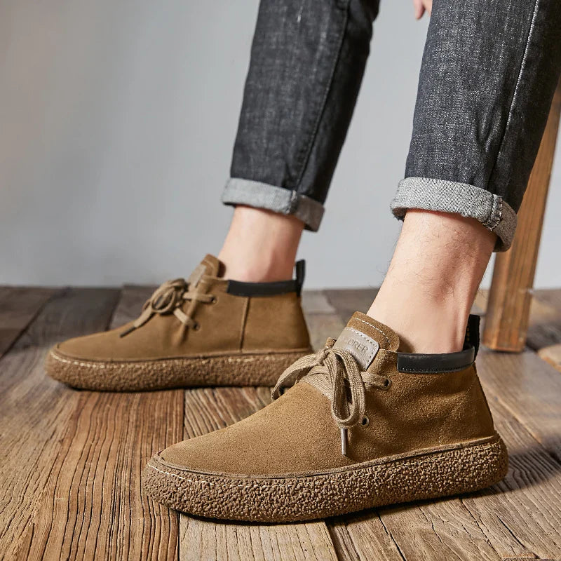 A person wearing rolled-up dark jeans and tan cow suede leather Oxfords dress shoes with textured brown rubber soles stands on wooden flooring.