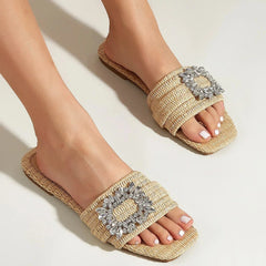 A pair of feet wearing handwoven cane slide sandals with square crystal buckles, shown from above on a light background. The toes have white nail polish.