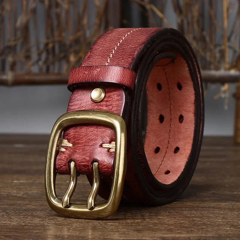 A genuine leather belt with visible stitching and a copper buckle is rolled up on a wooden surface. The belt showcases heritage craftsmanship, featuring multiple adjustment holes for versatility.