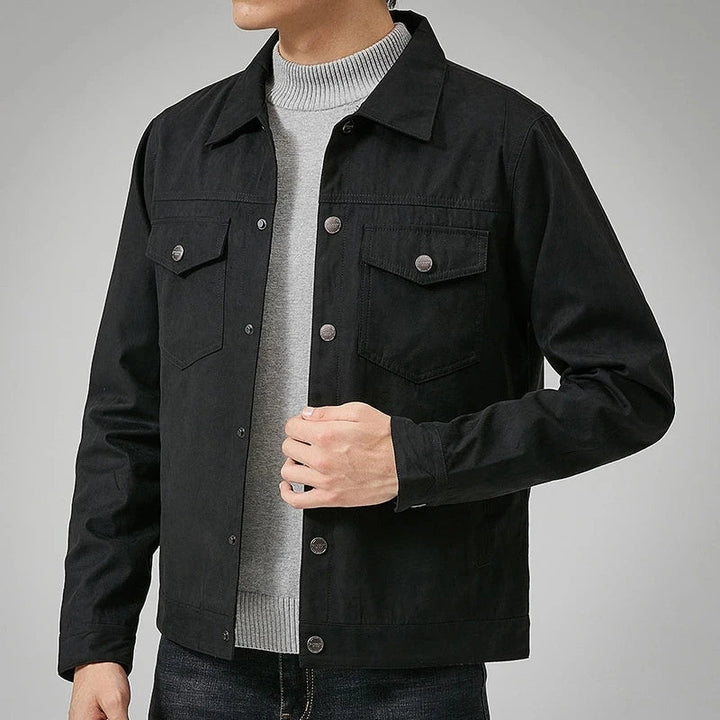 A person wears a black button-up jacket with a classic point collar over a light gray sweater and dark jeans, one hand adjusting the jacket. The face is not visible.