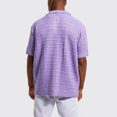 A person stands facing away from the camera, wearing a light purple, short-sleeved summer shirt with a breathable knit design and white pants against a plain light background.