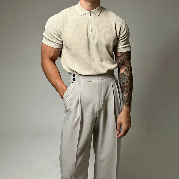 A man with a muscular build wears a beige quarter zip polo with ribbed detailing and high-waisted, pleated beige trousers. He stands with one hand in his pocket, showing off a tattoo sleeve on his left arm.