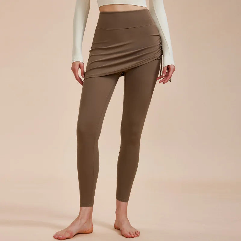 A person wearing high-waisted leggings with a matching asymmetrical skirt overlay and a long-sleeve cream top, barefoot against a neutral background—an effortless look for your autumn wardrobe.