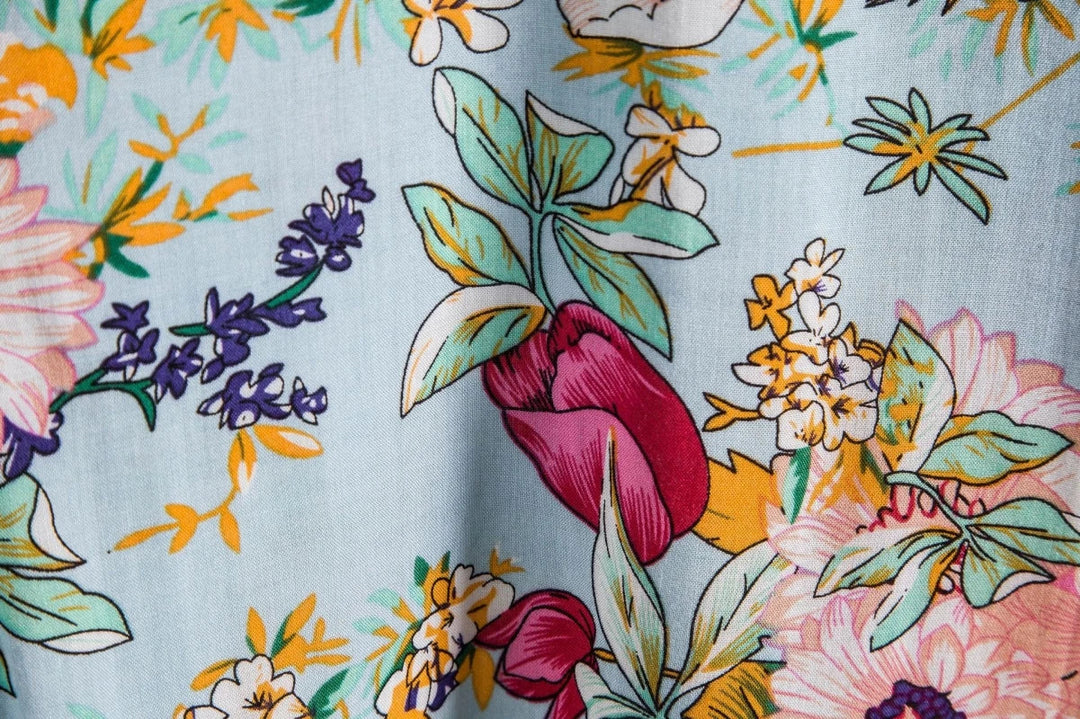 A close-up of colorful floral fabric, perfect for a luxury linen shirt, featuring large flowers, leaves, and stems in pink, yellow, green, and purple on a light blue background for breathable comfort.