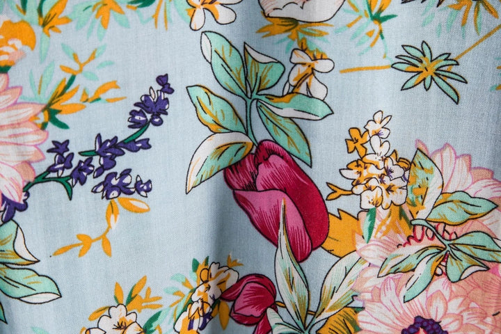 A close-up of colorful floral fabric, perfect for a luxury linen shirt, featuring large flowers, leaves, and stems in pink, yellow, green, and purple on a light blue background for breathable comfort.