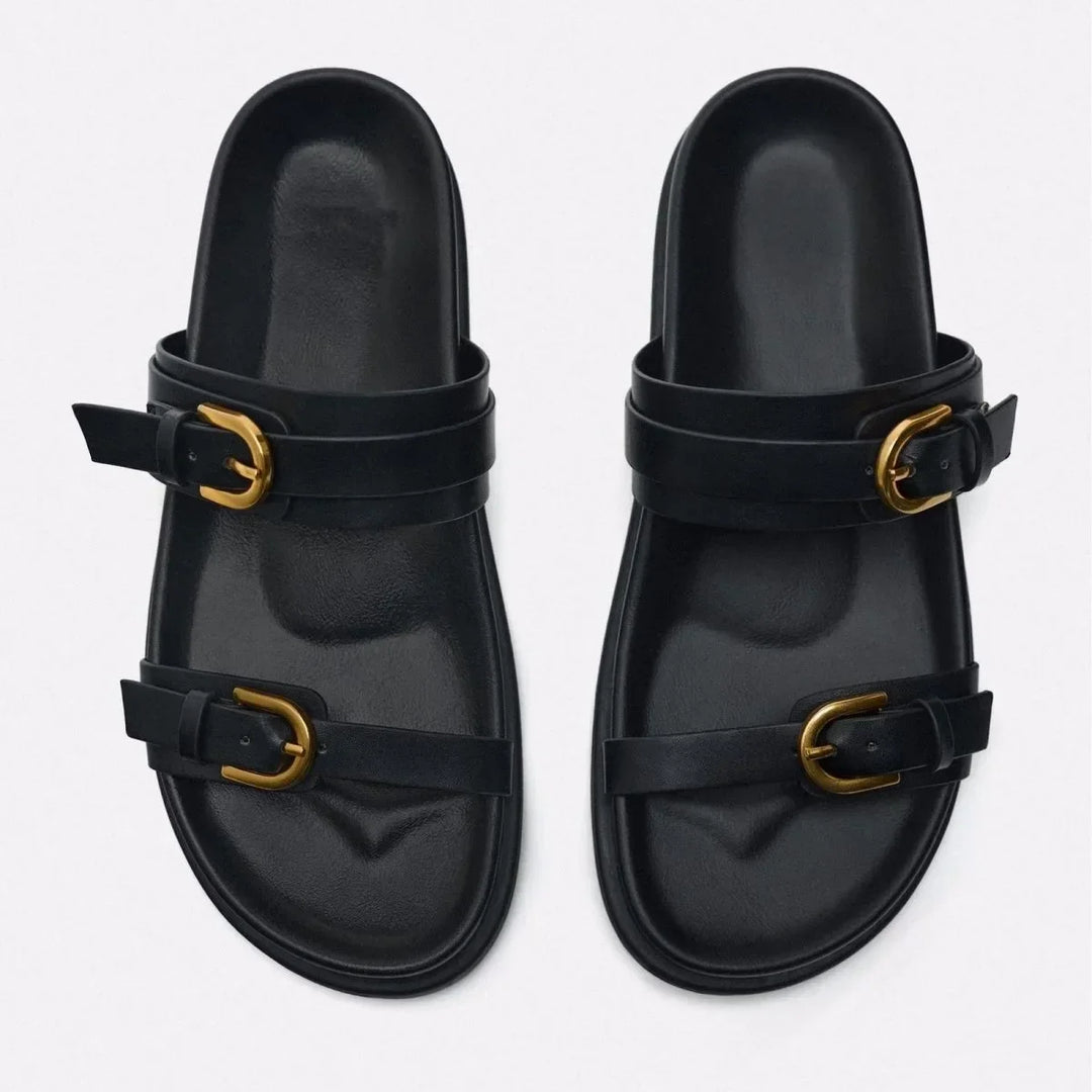 A pair of black slide sandals with a leather upper and two straps on each, featuring gold buckles and an ergonomic footbed, displayed on a light background.