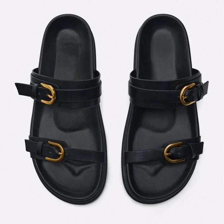 A pair of black slide sandals with a leather upper and two straps on each, featuring gold buckles and an ergonomic footbed, displayed on a light background.
