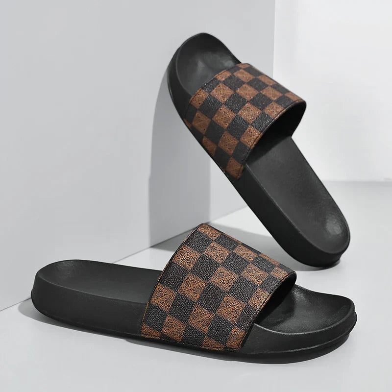A pair of black streetwear slides with wide straps featuring a brown and black checkerboard pattern, displayed against a white and gray background.