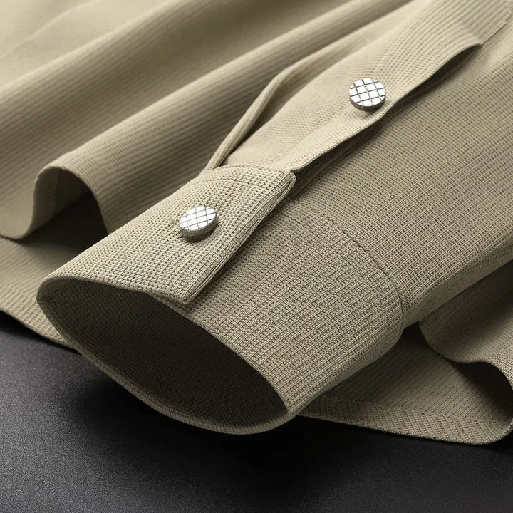 Close-up of a beige, wrinkle-resistant shirt cuff with two silver, grid-patterned buttons on a dark surface. The fabric has a fine, ribbed appearance, highlighting the premium quality of this anti-wrinkle dress shirt.