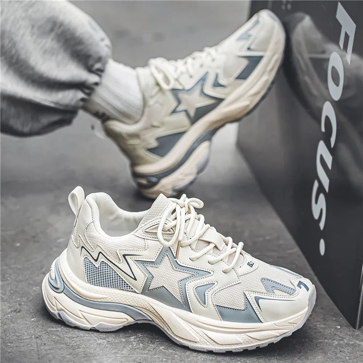 A person wearing white and light gray chunky sneakers with star patterns and a durable rubber outsole sits on the ground, one foot propped against a black box labeled Focus. They are dressed in light gray sweatpants.