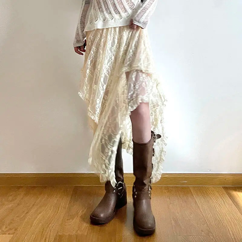 Person wearing a sheer white long-sleeve top, a silky lace skirt with an asymmetrical hemline and high waist, paired with brown knee-high boots, standing indoors on a wooden floor against a plain white wall.
