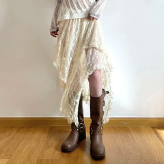 Person wearing a sheer white long-sleeve top, a silky lace skirt with an asymmetrical hemline and high waist, paired with brown knee-high boots, standing indoors on a wooden floor against a plain white wall.