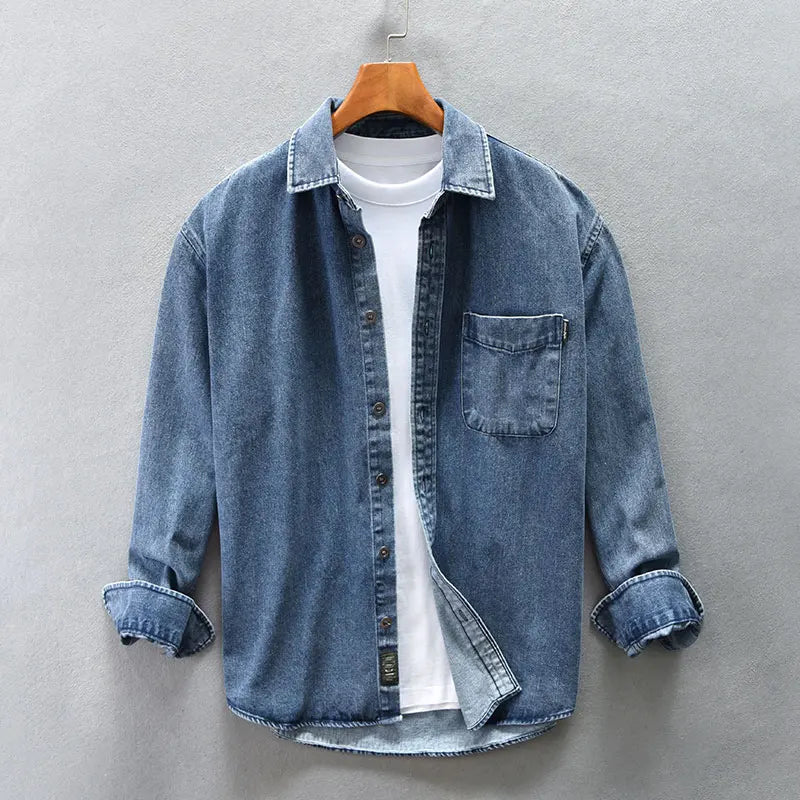 A blue washed denim overshirt with rolled-up sleeves is layered over a plain white t-shirt. Crafted from natural cotton, it features a front chest pocket and is displayed on a wooden hanger against a gray background.