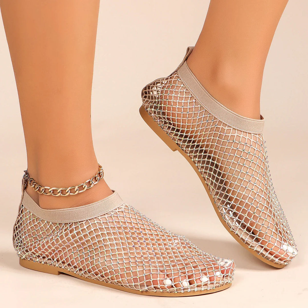 A pair of feet wearing beige slip-on mesh overlay shoes with a silver mesh design and cushioned footbed, exposing white-painted toenails. One ankle has a gold chain anklet. The background is a neutral beige color.
