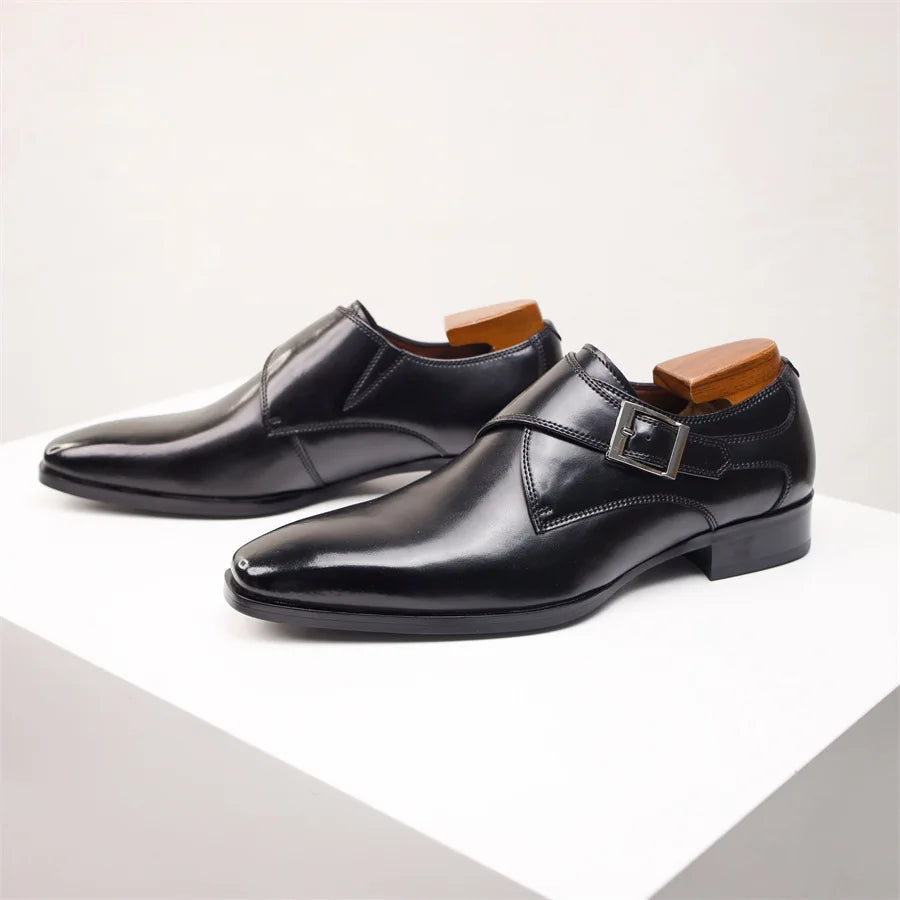 A pair of shiny black Italian leather monk strap shoes with a side buckle detail, displayed on a white surface. Shoe trees are inserted to maintain their shape.