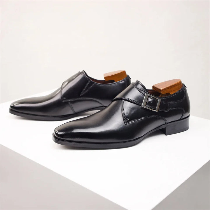 A pair of shiny black Italian leather monk strap shoes with a side buckle detail, displayed on a white surface. Shoe trees are inserted to maintain their shape.