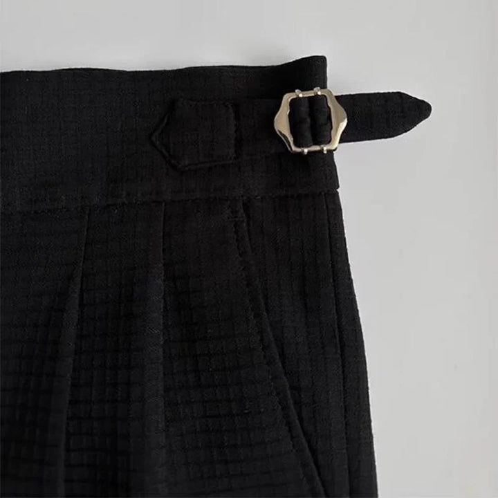 Close-up of the waistband of black luxury tailored pants with a silver buckle and strap detail on the side, set against a light background.