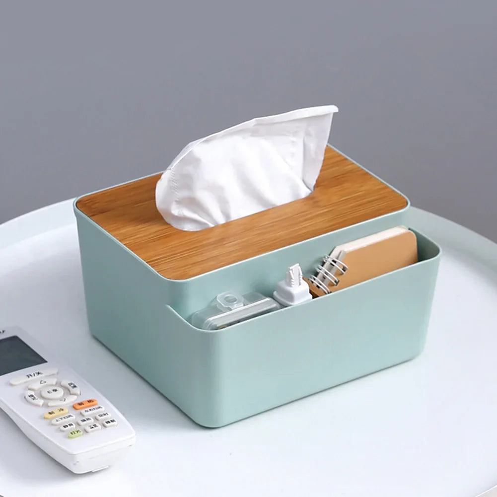 A mint green Table Wooden Cover Tissue Box with a wooden lid acts as a desktop organizer, holding tissues, a small notebook, a phone charger, and a remote control on a white surface.