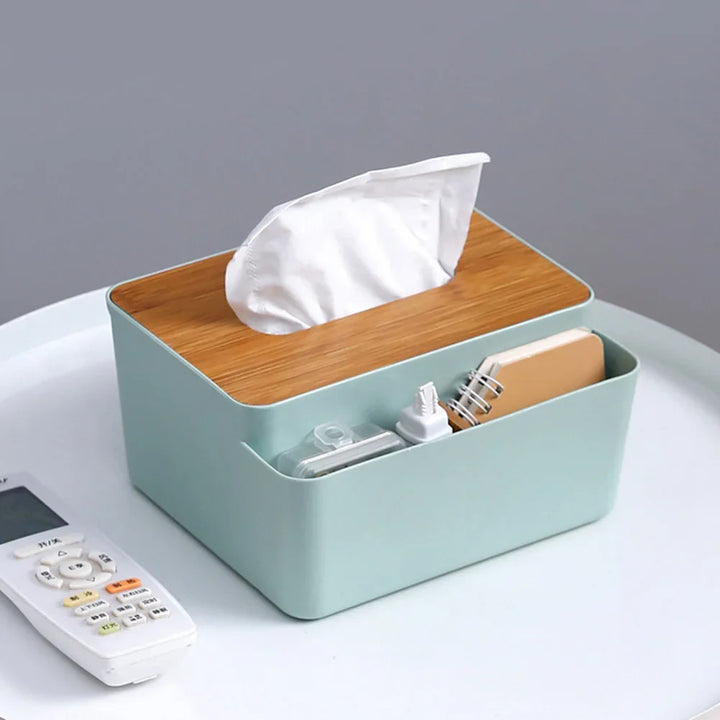 A mint green Table Wooden Cover Tissue Box with a wooden lid acts as a desktop organizer, holding tissues, a small notebook, a phone charger, and a remote control on a white surface.