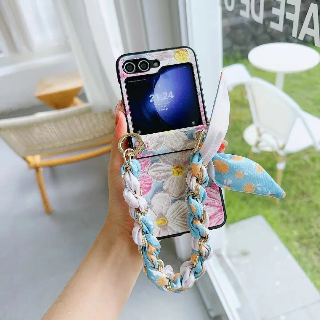 A hand holds a foldable smartphone with a floral case, a pastel braided chain strap, and a blue polka dot scarf. The slim design fits perfectly in hand. A chair and round table are seen in the bright, airy background as the phone displays the time.