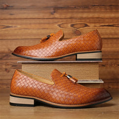 A pair of brown real leather loafers with tassels, featuring a basketweave pattern, is displayed against a wooden background. One shoe sits atop two stacked books, while the other rests on the surface below.