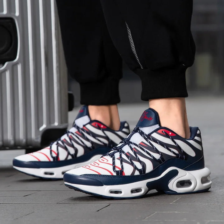 A person wearing stylish navy blue, white, and red athletic sneakers with a cushioned midsole and black pants stands on a sidewalk next to a silver suitcase.