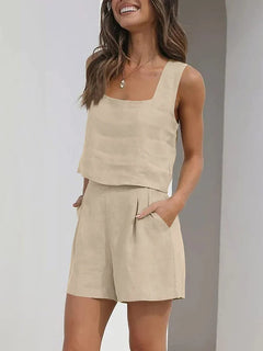 Woman standing and smiling, wearing a contemporary style, light beige linen set with a square-neck sleeveless vest and matching high-waisted shorts, her hands casually placed in the pockets.
