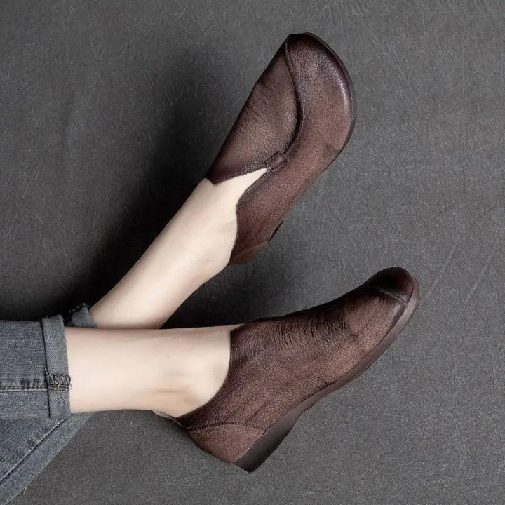 A person wearing dark brown, ankle-high shoes crafted from supple leather with a slight wedge heel and gray rolled-up jeans, against a textured gray background.