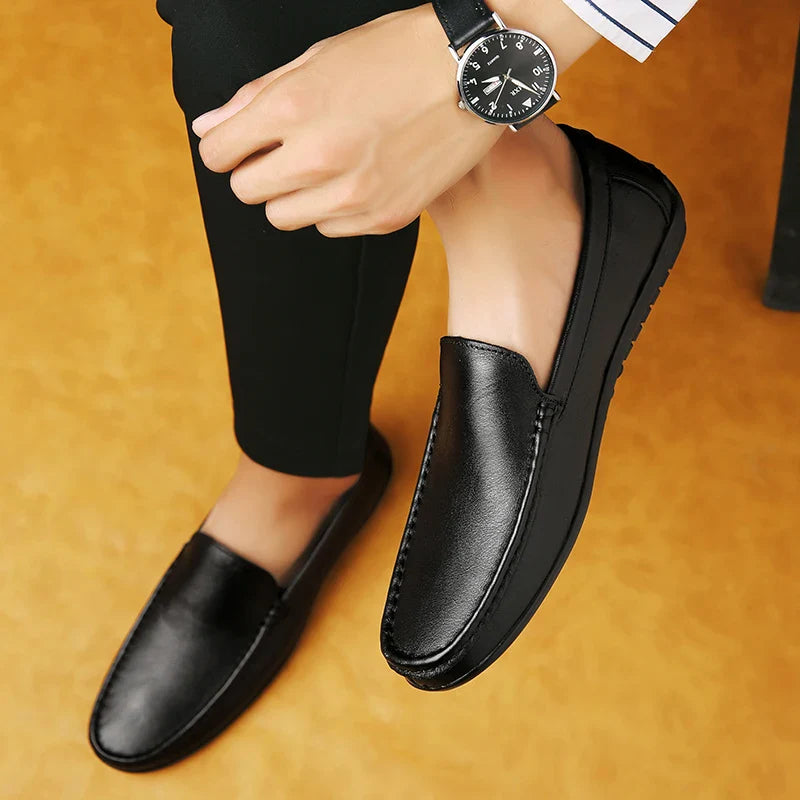A person wearing luxury shoes with a cushioned footbed and a black wristwatch sits with legs crossed on a yellowish-brown floor, dressed in black pants and a shirt featuring white and blue striped cuffs.